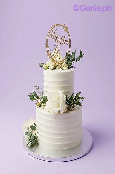 Wedding White Floral 2-Tier Cake White 2-tier wedding cake with textured soft icing, a Mr & Mrs circular topper, and fresh white roses with greenery.