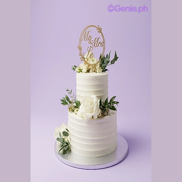Wedding White Floral 2-Tier Cake White 2-tier wedding cake with textured soft icing, a Mr & Mrs circular topper, and fresh white roses with greenery.