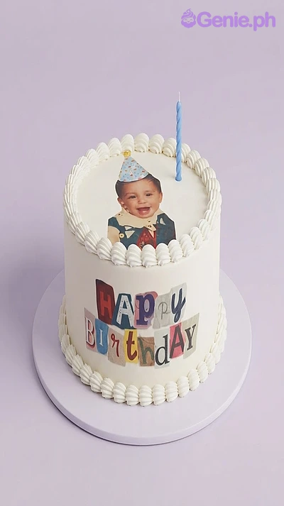 White 1-tier cake with soft icing, an edible photo of a child on top, a blue candle, and a Happy Birthday banner on the side