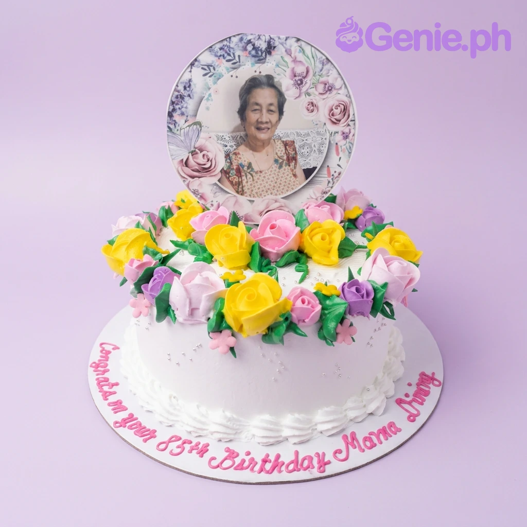 85th Birthday White Floral Cake A white 1-tier birthday cake themed for an 85th birthday, featuring a photo printout and a wreath of pink, yellow, and purple icing roses.