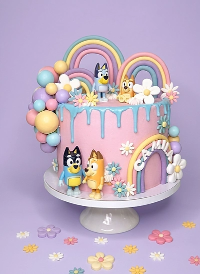 Pink Bluey themed birthday cake with fondant rainbows, 3D character figures, blue icing drip, and colorful gumpaste flowers. Pink Bluey themed birthday cake with fondant rainbows, 3D character figures, blue icing drip, and colorful gumpaste flowers.