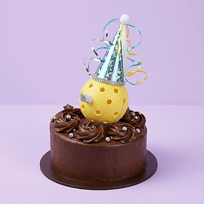 Chocolate cake with dark brown icing swirls, silver dragees, and a yellow ball topper wearing a blue party hat.