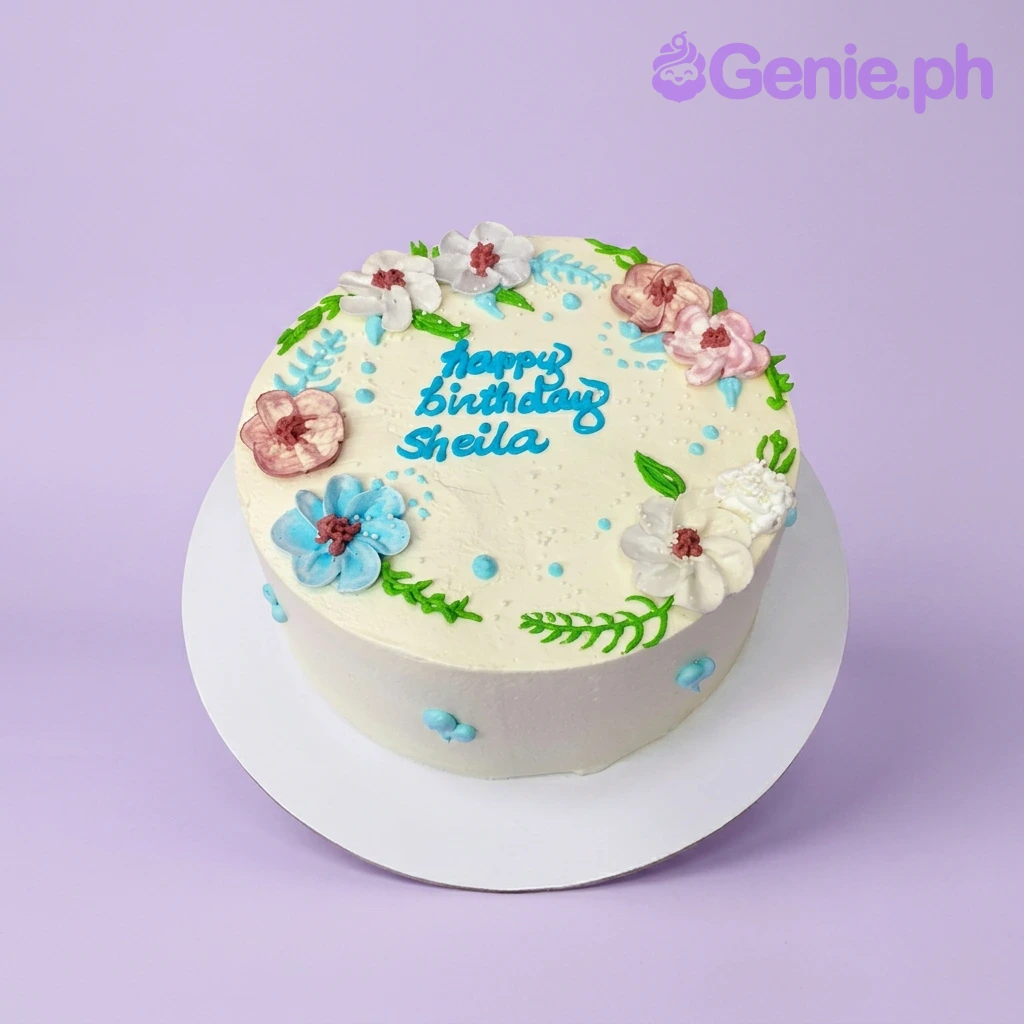Floral Birthday White Cake A white 1-tier cake themed around Floral Birthday, featuring colorful piped icing flowers and a blue 'Happy Birthday Sheila' message.