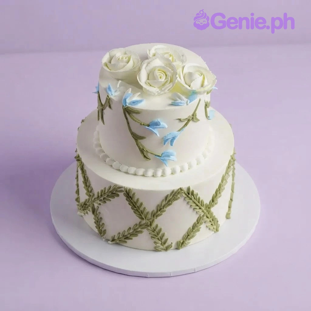 Floral Lattice Ivory 2-Tier Cake Ivory 2-tier soft icing cake with piped white roses, blue floral vines, and a green leaf lattice pattern on the bottom tier