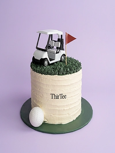 White textured icing golf themed cake with a white golf cart topper, red flag, and a large golf ball on a dark green base.