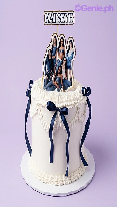 Katseye-Inspired Kpop White Birthday Cake White soft-icing cake with navy blue ribbons and a Katseye K-pop group printout topper.