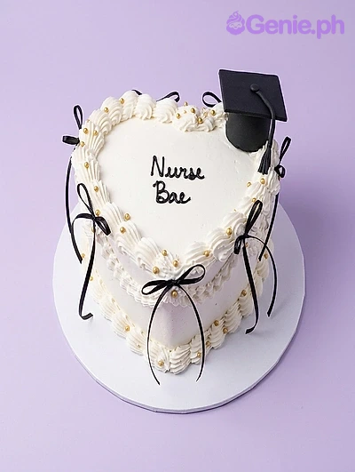 Nurse Graduation White Heart Cake A white heart-shaped cake for Nurse Graduation, featuring a black graduation cap, white ruffle icing, black bows, and gold pearls.