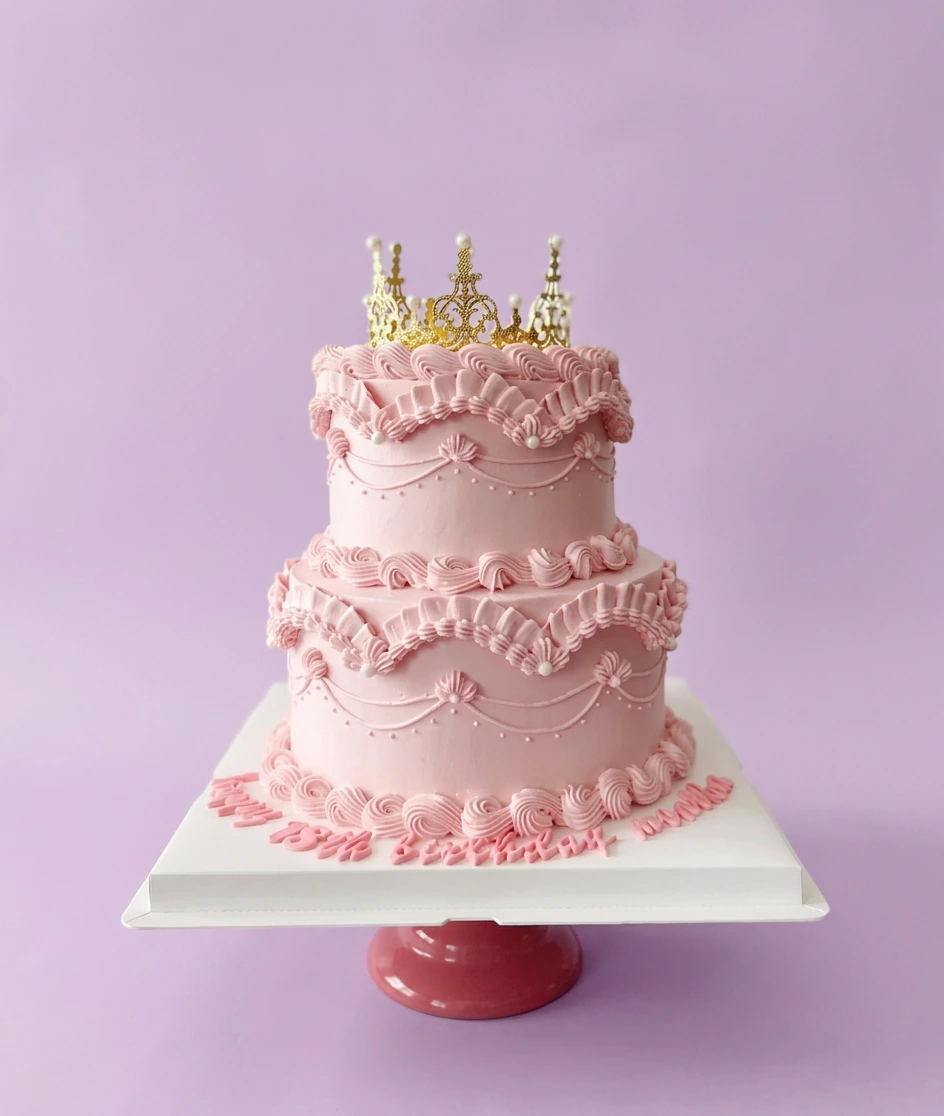Elegant 2-tier light pink vintage lambeth cake with intricate ruffles, white pearls, and a gold crown topper.