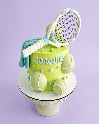 Light green tennis themed cake with a large fondant tennis racket and balls, featuring the name Joaquin in blue letters.
