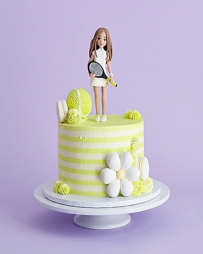 Tennis themed birthday cake with a girl tennis player figurine, tennis ball, and white flower on a lime green striped base.
