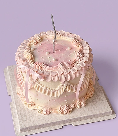 Vintage-style pink and white ruffled cake with delicate icing swags, pink ribbons, and a single tall silver candle.