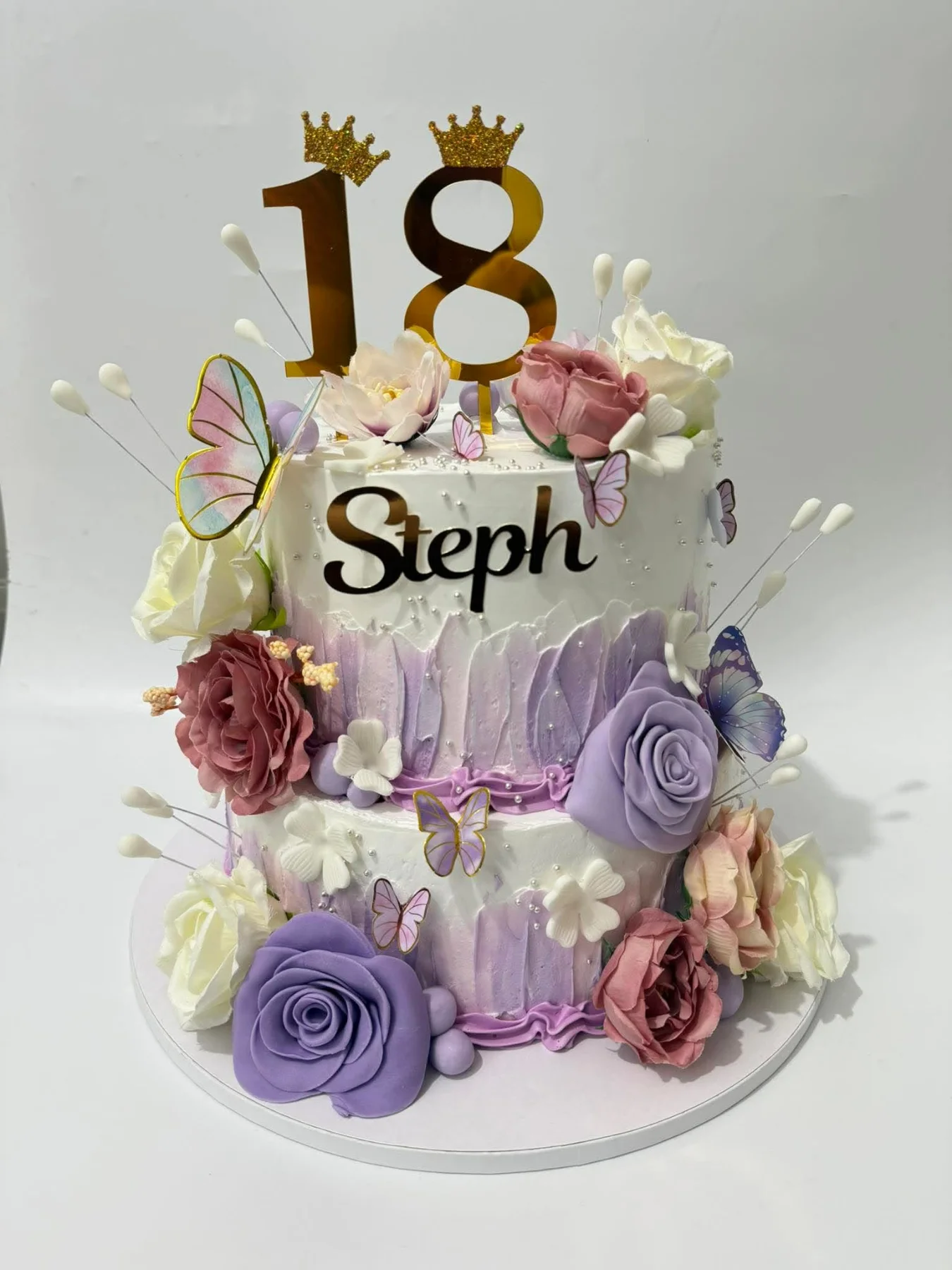 Elegant 2-tier white and purple cake with palette knife icing, edible roses, butterflies, and a gold number 18 topper. Elegant 2-tier white and purple cake with palette knife icing, edible roses, butterflies, and a gold number 18 topper.