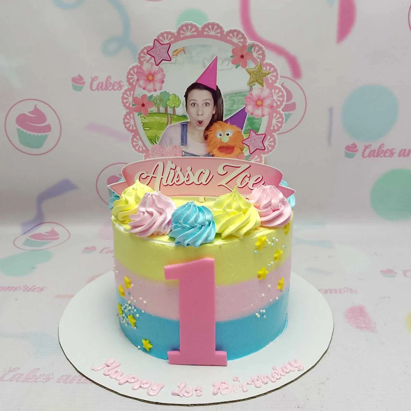 A colorful 1st birthday cake with yellow, pink, and blue horizontal icing stripes, topped with a custom photo printout and icing swirls. A colorful 1st birthday cake with yellow, pink, and blue horizontal icing stripes, topped with a custom photo printout and icing swirls.
