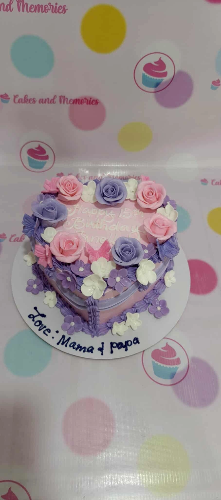 Heart-shaped pink cake decorated with purple and pink fondant roses and white flowers.