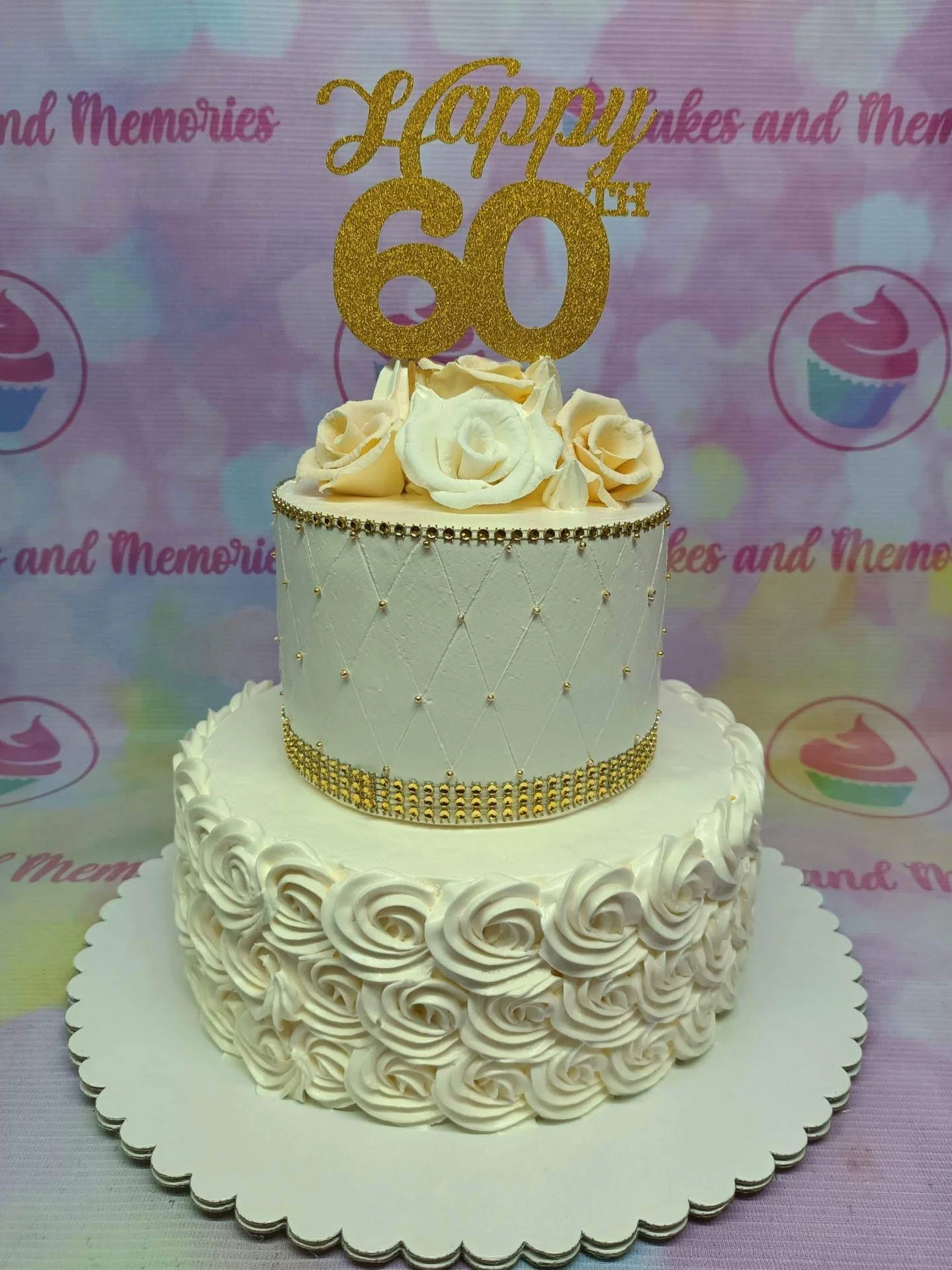 60th Birthday White Floral 2-Tier Cake A white 2-tier 60th Birthday cake with gold glitter 'Happy 60th' topper, cream roses, quilted icing, and gold beads.