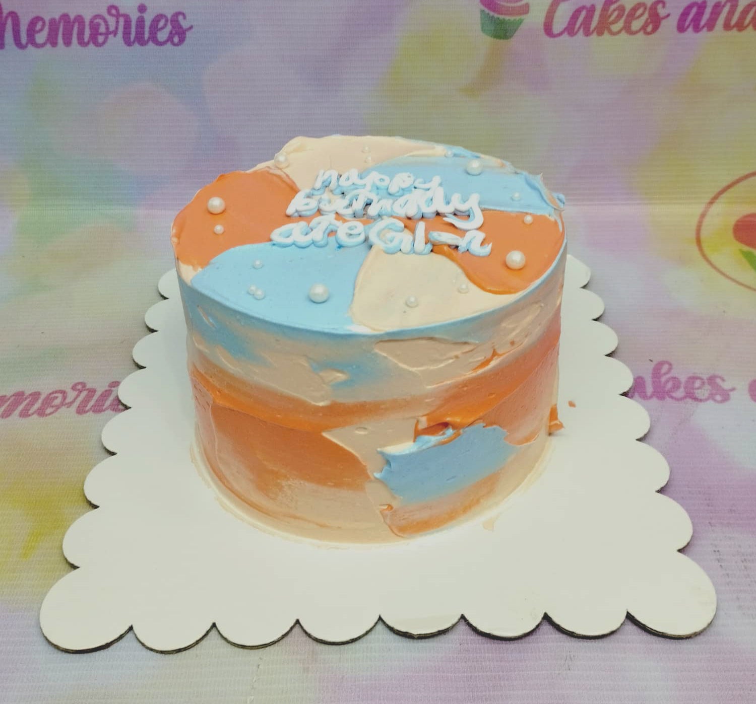 A minimalist 1-tier birthday cake with abstract blue, orange, and peach palette knife icing, topped with white pearls and a piped message.