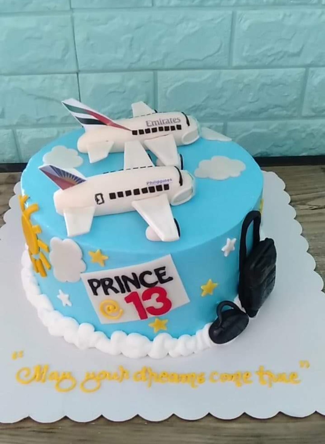 Light blue airplane themed birthday cake with Emirates and Philippines airline toppers, white clouds, and black luggage decorations. Light blue airplane themed birthday cake with Emirates and Philippines airline toppers, white clouds, and black luggage decorations.