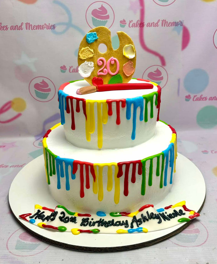 Two-tier white cake with colorful paint drips, a gold artist palette topper, and a paintbrush decoration. Two-tier white cake with colorful paint drips, a gold artist palette topper, and a paintbrush decoration.