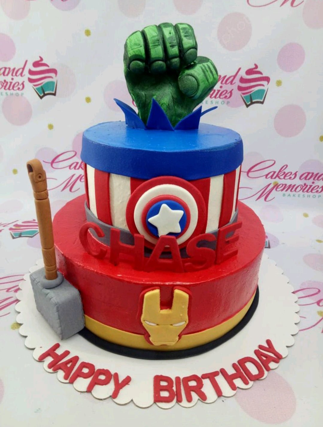 Two-tier Avengers themed cake featuring Hulk's fist, Captain America's shield, Iron Man's mask, and Thor's hammer. Two-tier Avengers themed cake featuring Hulk's fist, Captain America's shield, Iron Man's mask, and Thor's hammer.