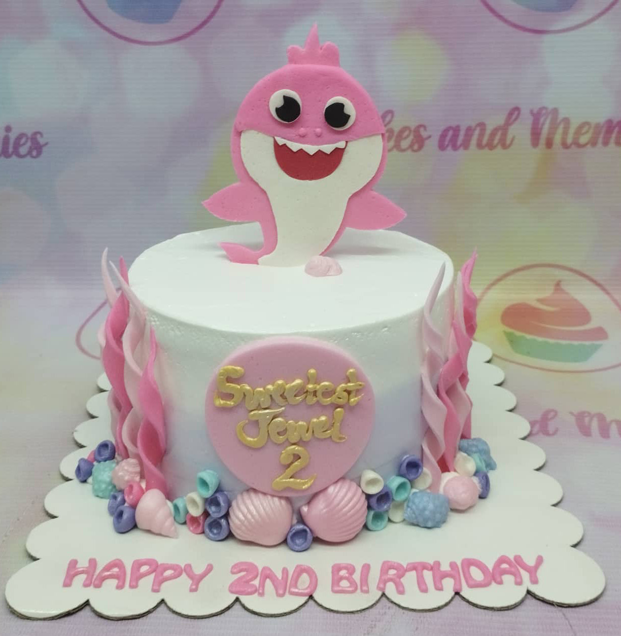 Pink Baby Shark themed birthday cake with fondant sea creatures and a pink shark topper on a white icing base. Pink Baby Shark themed birthday cake with fondant sea creatures and a pink shark topper on a white icing base.