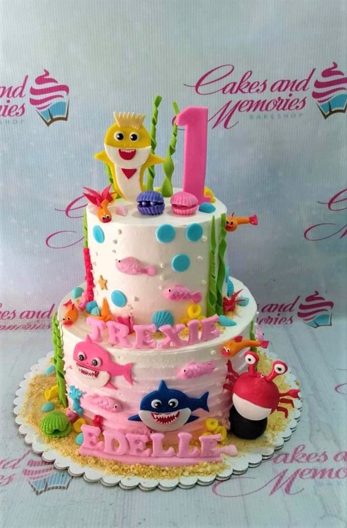 Two-tier Baby Shark themed birthday cake with pink and blue icing, fondant sea creatures, and a yellow shark topper. Two-tier Baby Shark themed birthday cake with pink and blue icing, fondant sea creatures, and a yellow shark topper.
