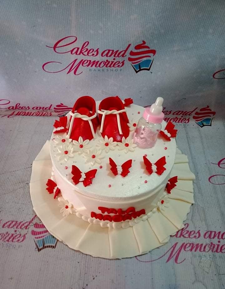 White round baby shower cake with red fondant shoes, a pink baby bottle, and red butterfly decorations. White round baby shower cake with red fondant shoes, a pink baby bottle, and red butterfly decorations.