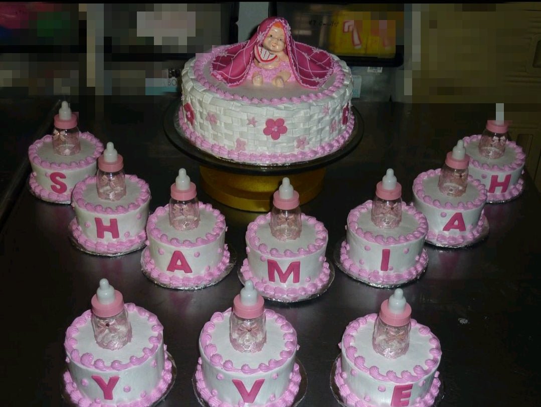 White basketweave cake with pink icing rosettes and a baby figurine topper surrounded by matching mini cakes with letters. White basketweave cake with pink icing rosettes and a baby figurine topper surrounded by matching mini cakes with letters.