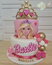 Pink and white 2-tier Barbie themed cake with a large gold crown, Barbie printout, and pink and gold decorative balls.