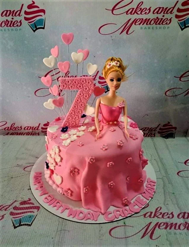 Pink Barbie themed birthday cake with a doll topper, pink heart toppers, and a large number 7 on a pink fondant base.