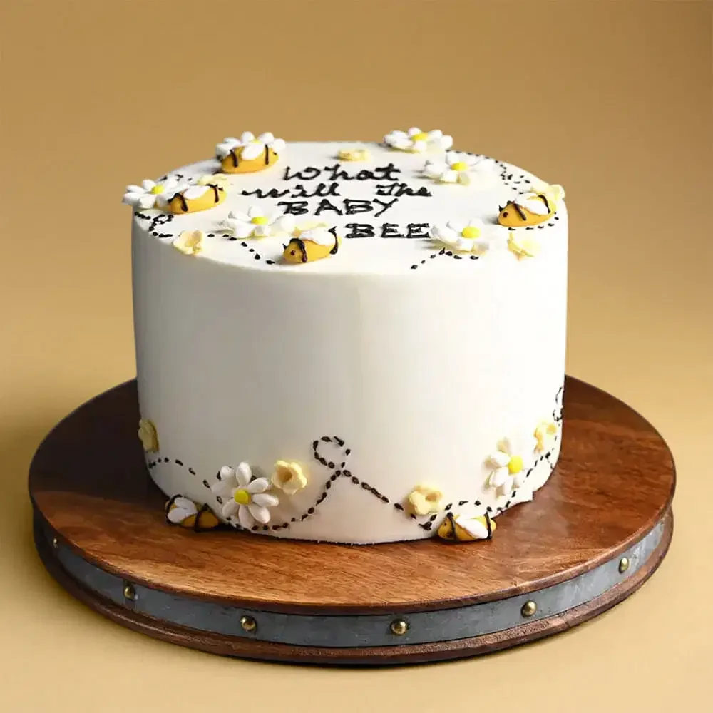 White 1-tier baby shower cake with yellow fondant bees, white daisies, and black piped icing trails on a wooden base. White 1-tier baby shower cake with yellow fondant bees, white daisies, and black piped icing trails on a wooden base.