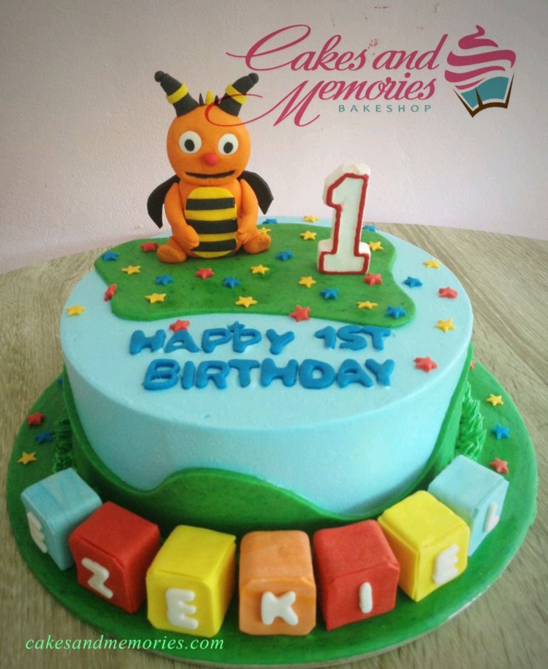 Light blue birthday cake with a 3D orange bee character, green grass icing, and colorful letter blocks spelling EZEKIEL. Light blue birthday cake with a 3D orange bee character, green grass icing, and colorful letter blocks spelling EZEKIEL.