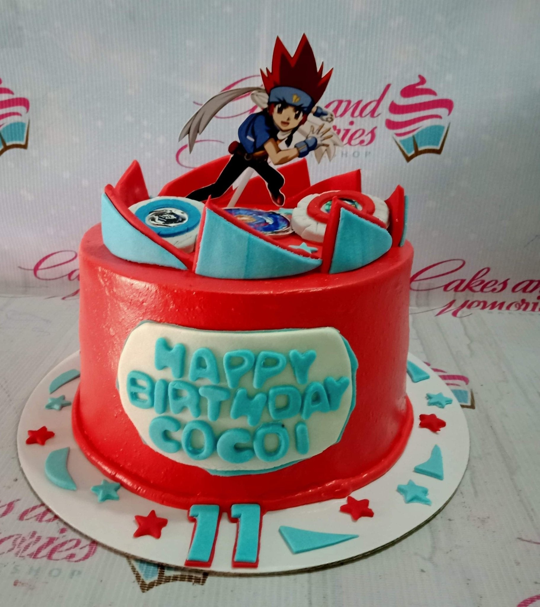 Red Beyblade themed birthday cake with a character printout topper and blue gumpaste accents. Red Beyblade themed birthday cake with a character printout topper and blue gumpaste accents.