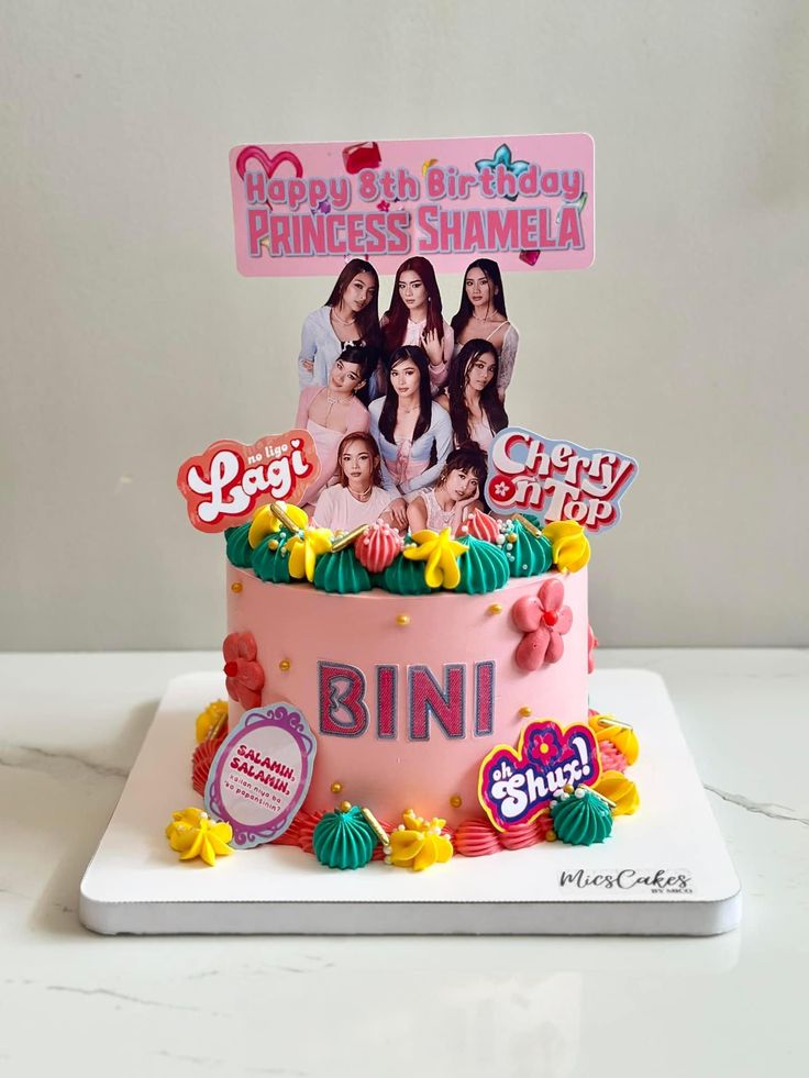 A pink 1 tier cake featuring a BINI group photo banner, song logo cutouts, icing swirls, fondant flowers, and gold sprinkles. A pink 1 tier cake featuring a BINI group photo banner, song logo cutouts, icing swirls, fondant flowers, and gold sprinkles.