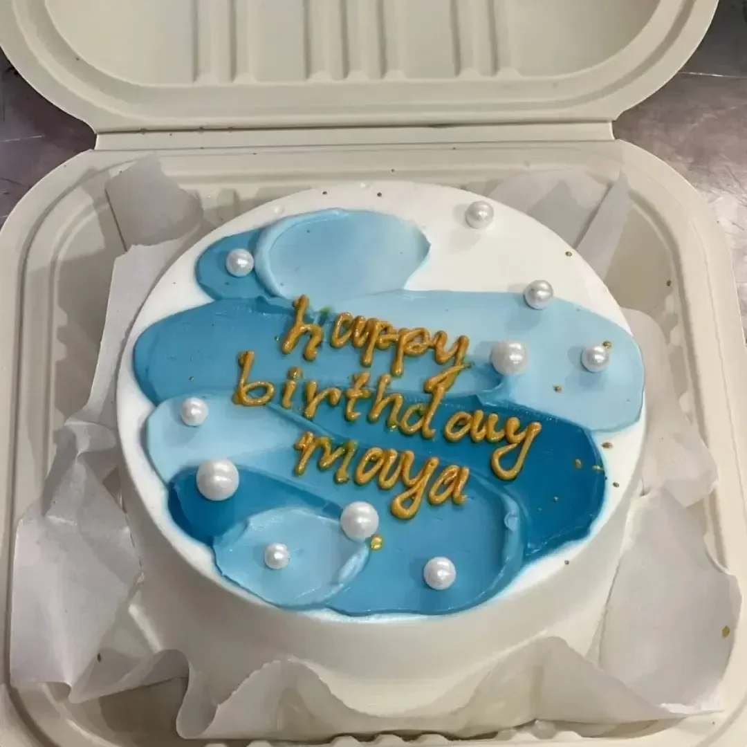 White bento cake with blue palette knife icing strokes, gold birthday message, and white pearls in a takeout container. White bento cake with blue palette knife icing strokes, gold birthday message, and white pearls in a takeout container.