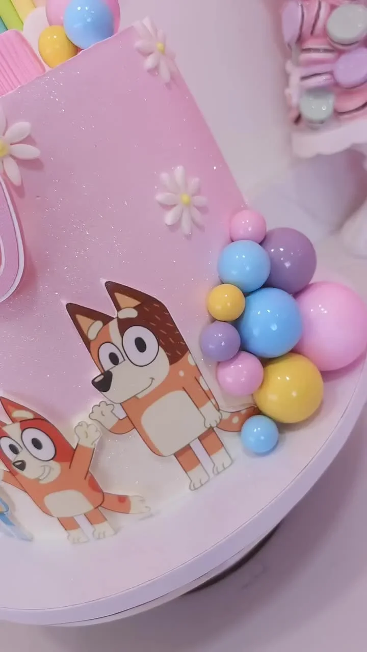 Pink Bluey themed birthday cake with glitter icing, character printouts, and colorful plastic ball decorations. Pink Bluey themed birthday cake with glitter icing, character printouts, and colorful plastic ball decorations.