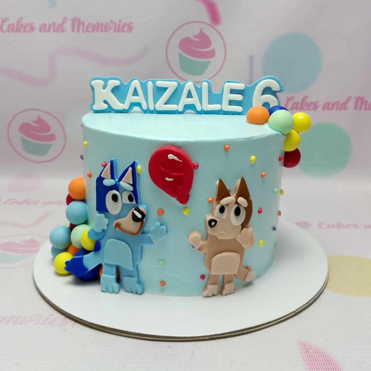 Light blue Bluey themed birthday cake with fondant characters Bluey and Bingo, a red balloon, and colorful fondant balls. Light blue Bluey themed birthday cake with fondant characters Bluey and Bingo, a red balloon, and colorful fondant balls.