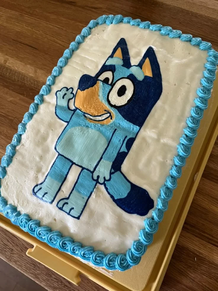 Rectangle Bluey character cake with white soft icing and blue piped borders. Rectangle Bluey character cake with white soft icing and blue piped borders.