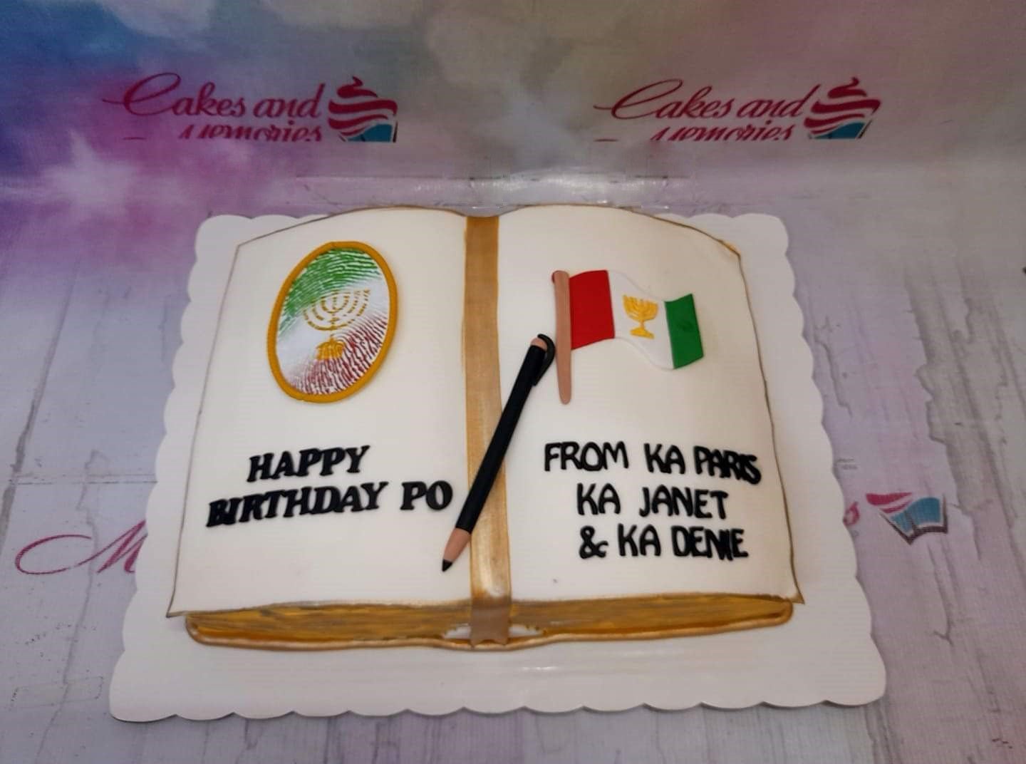 Open book shaped cake with white icing, gold edges, a black pencil, and religious symbols for a birthday celebration. Open book shaped cake with white icing, gold edges, a black pencil, and religious symbols for a birthday celebration.