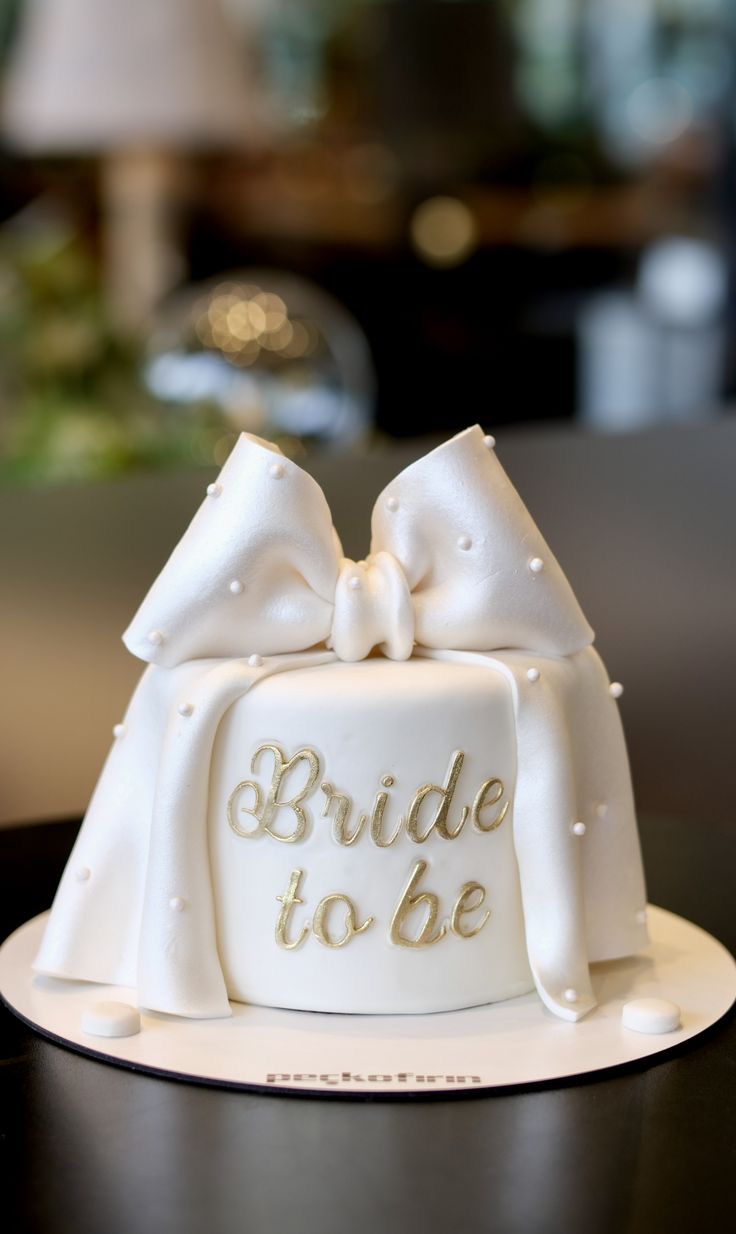 A white fondant cake featuring a large white bow with pearl accents, white pearl sprinkles, and a gold 'Bride to be' message. A white fondant cake featuring a large white bow with pearl accents, white pearl sprinkles, and a gold 'Bride to be' message.