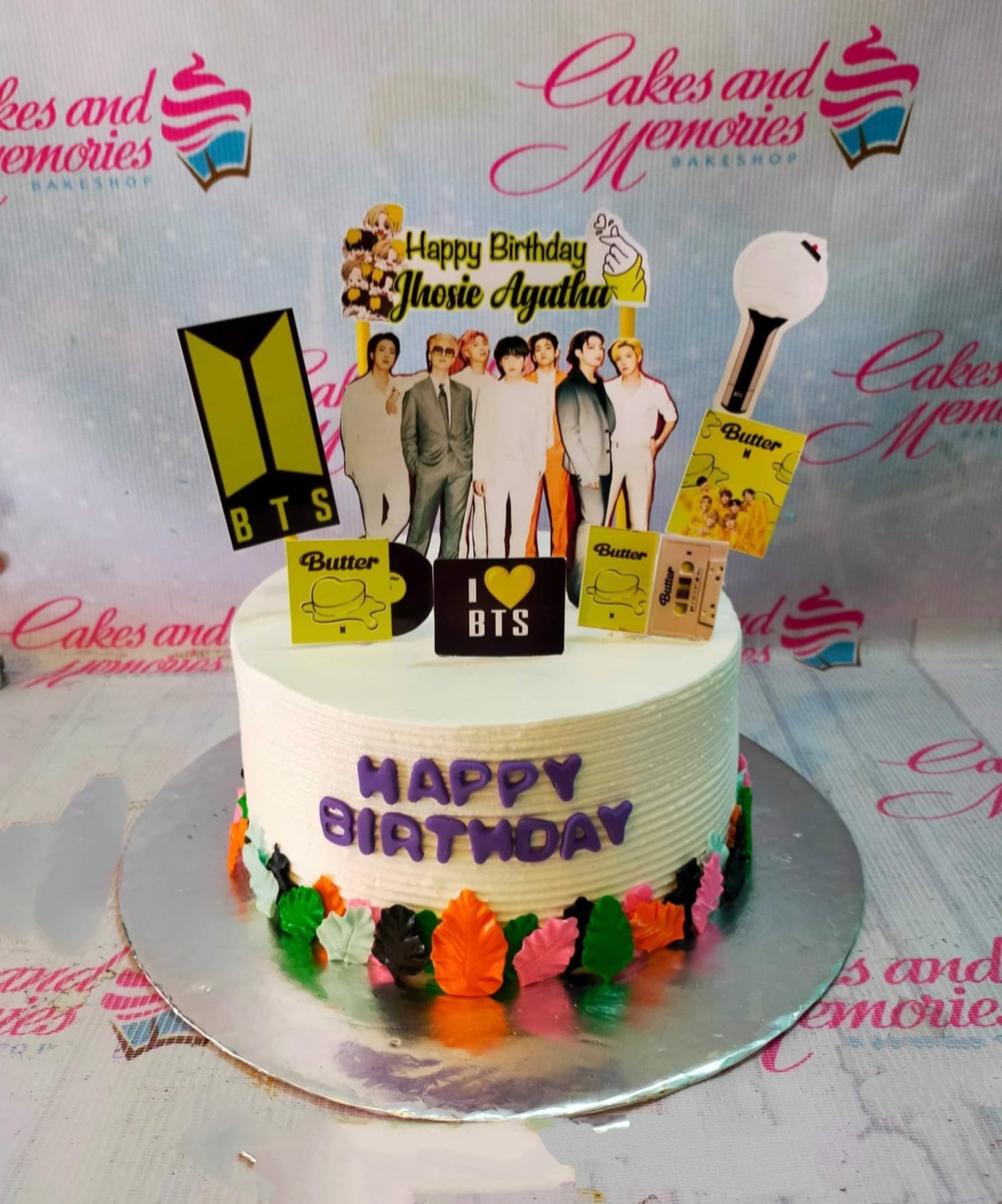 White BTS themed birthday cake with purple icing text and colorful fondant leaf border featuring various BTS member printouts. White BTS themed birthday cake with purple icing text and colorful fondant leaf border featuring various BTS member printouts.