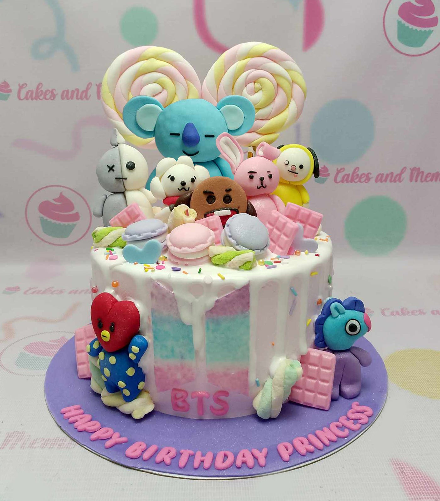BTS BT21 themed birthday cake with colorful fondant characters, pink chocolate bars, and rainbow lollipops on a purple base. BTS BT21 themed birthday cake with colorful fondant characters, pink chocolate bars, and rainbow lollipops on a purple base.