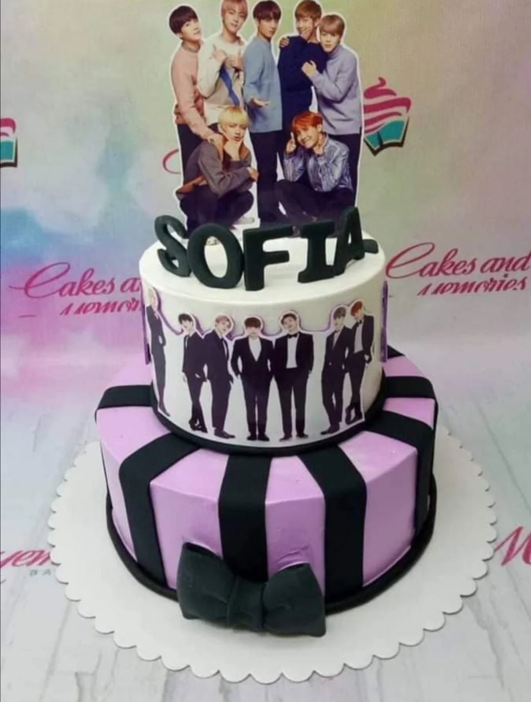 BTS Kpop-Inspired Bow 2-Tier Cake Two-tier purple and white BTS themed cake with black stripes, a large black bow, and printed character toppers.