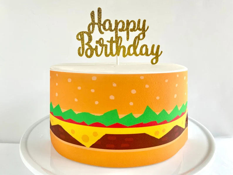 Single tier burger themed birthday cake with a gold glitter Happy Birthday topper and edible burger wrap design. Single tier burger themed birthday cake with a gold glitter Happy Birthday topper and edible burger wrap design.