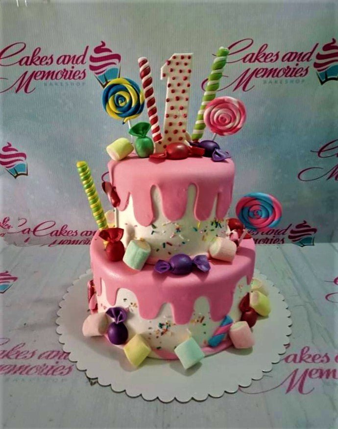 Two-tier pink and white candy-themed birthday cake with lollipops, marshmallows, and a number one topper. Two-tier pink and white candy-themed birthday cake with lollipops, marshmallows, and a number one topper.