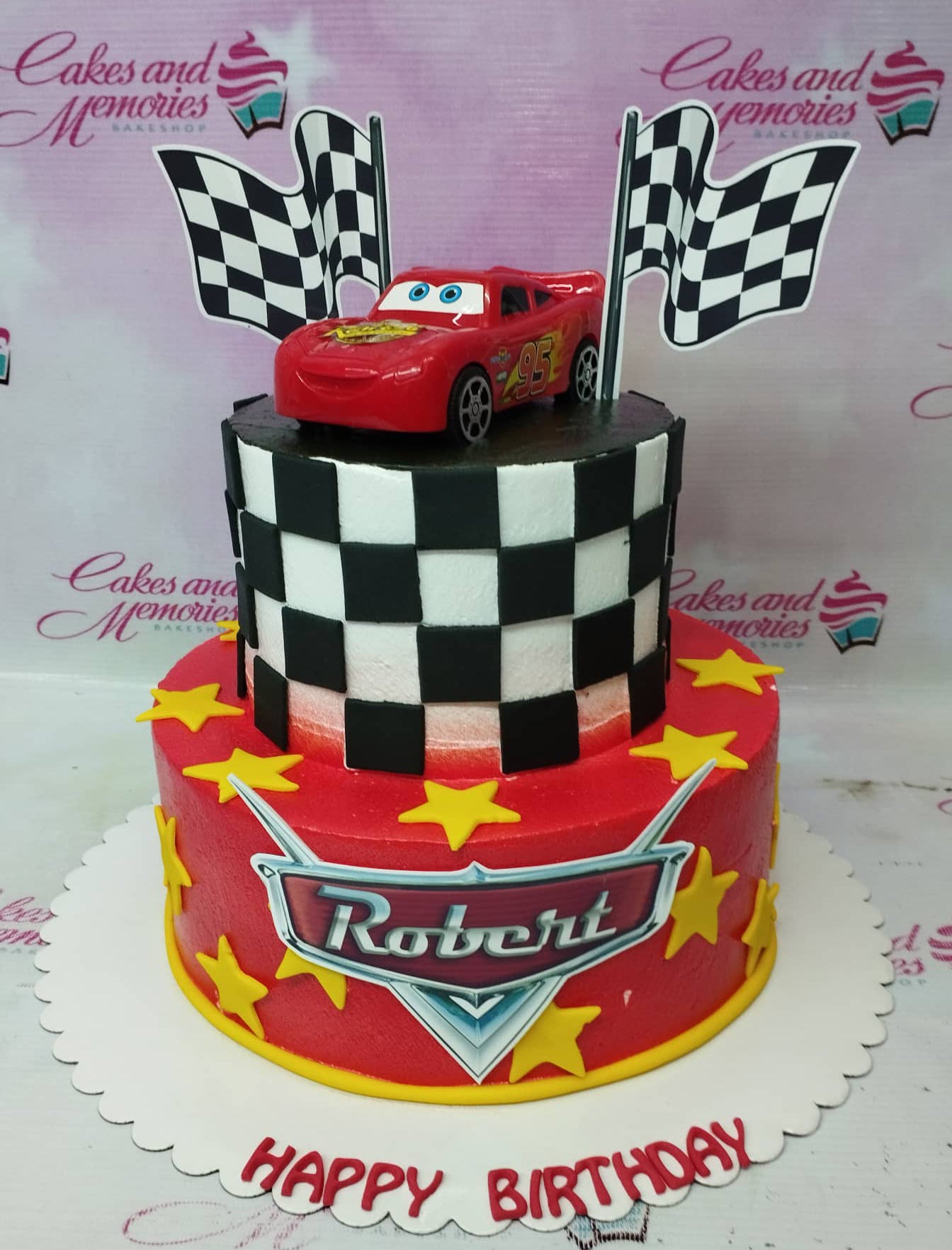 Two-tier Disney Cars themed birthday cake with Lightning McQueen toy topper, checkered flag printouts, and yellow fondant stars on red icing. Two-tier Disney Cars themed birthday cake with Lightning McQueen toy topper, checkered flag printouts, and yellow fondant stars on red icing.