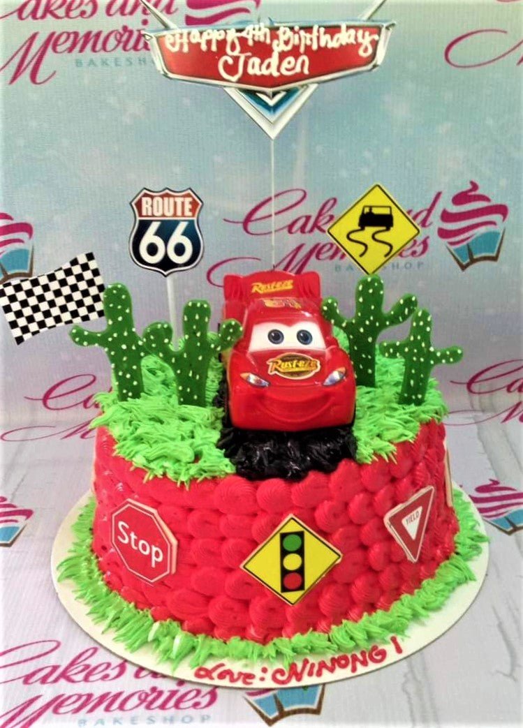 Red Cars themed birthday cake with Lightning McQueen toy topper, cactus decorations, and road signs on green grass icing. Red Cars themed birthday cake with Lightning McQueen toy topper, cactus decorations, and road signs on green grass icing.