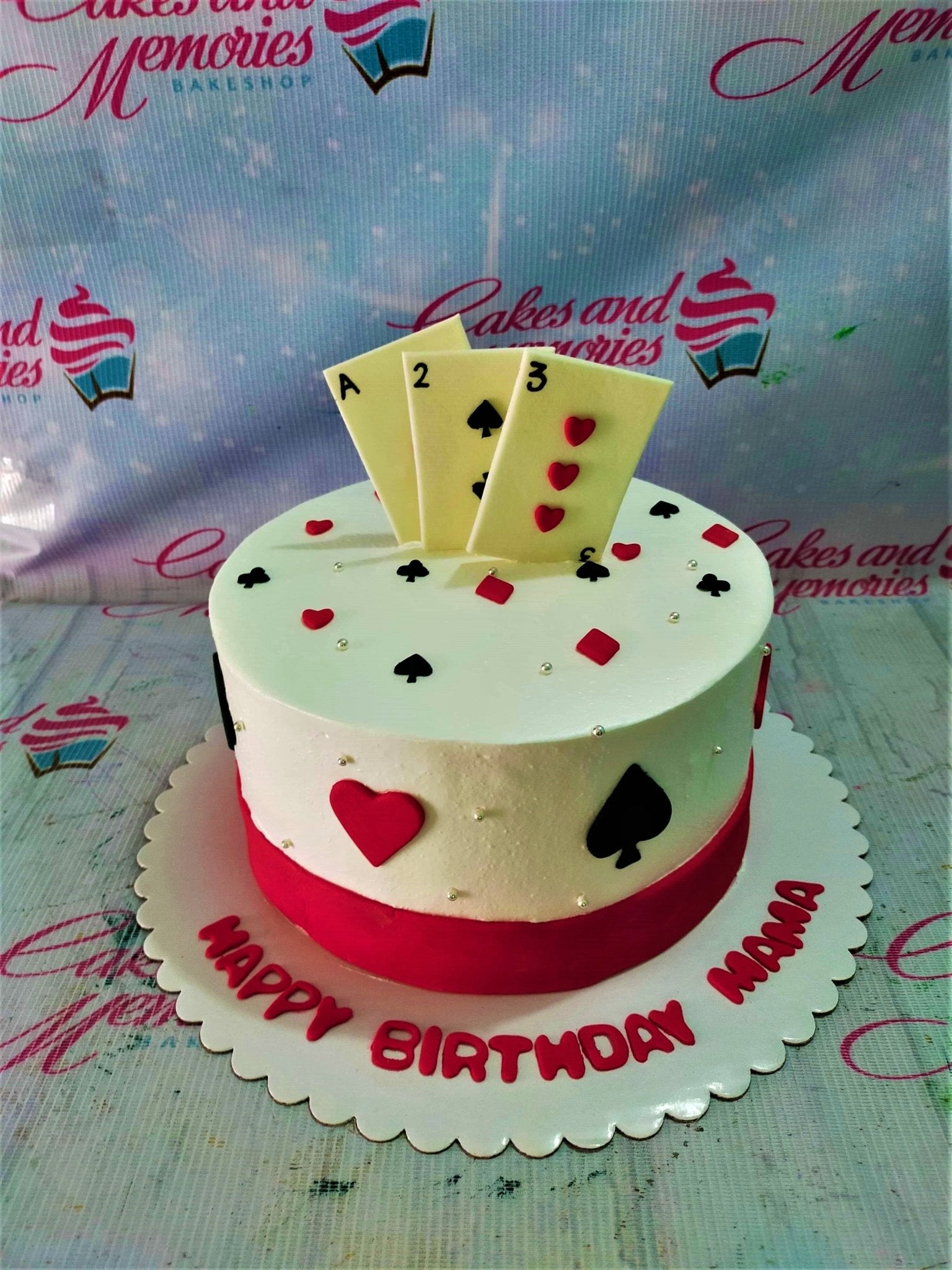 White round cake with playing card toppers, red and black card suit decorations, and a red base border. White round cake with playing card toppers, red and black card suit decorations, and a red base border.