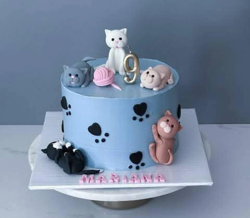 Light blue cat themed birthday cake with multiple 3D fondant cats, a pink yarn ball, and black paw prints on the side. Light blue cat themed birthday cake with multiple 3D fondant cats, a pink yarn ball, and black paw prints on the side.