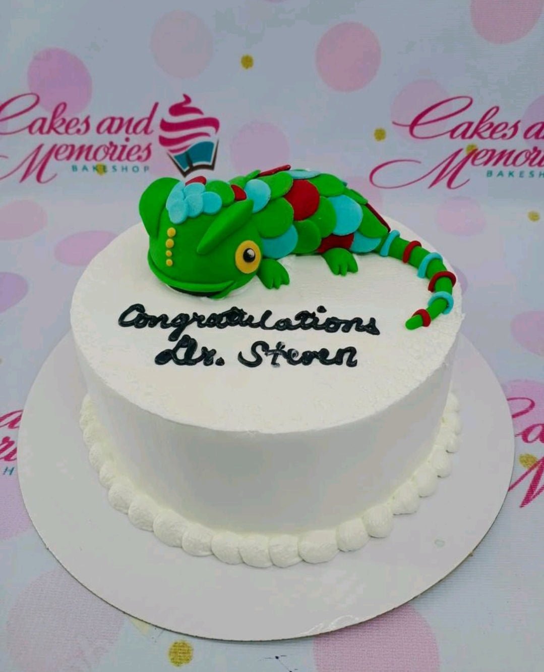 White round cake with a large green 3D fondant chameleon topper and black piped icing message for Dr. Steven. White round cake with a large green 3D fondant chameleon topper and black piped icing message for Dr. Steven.