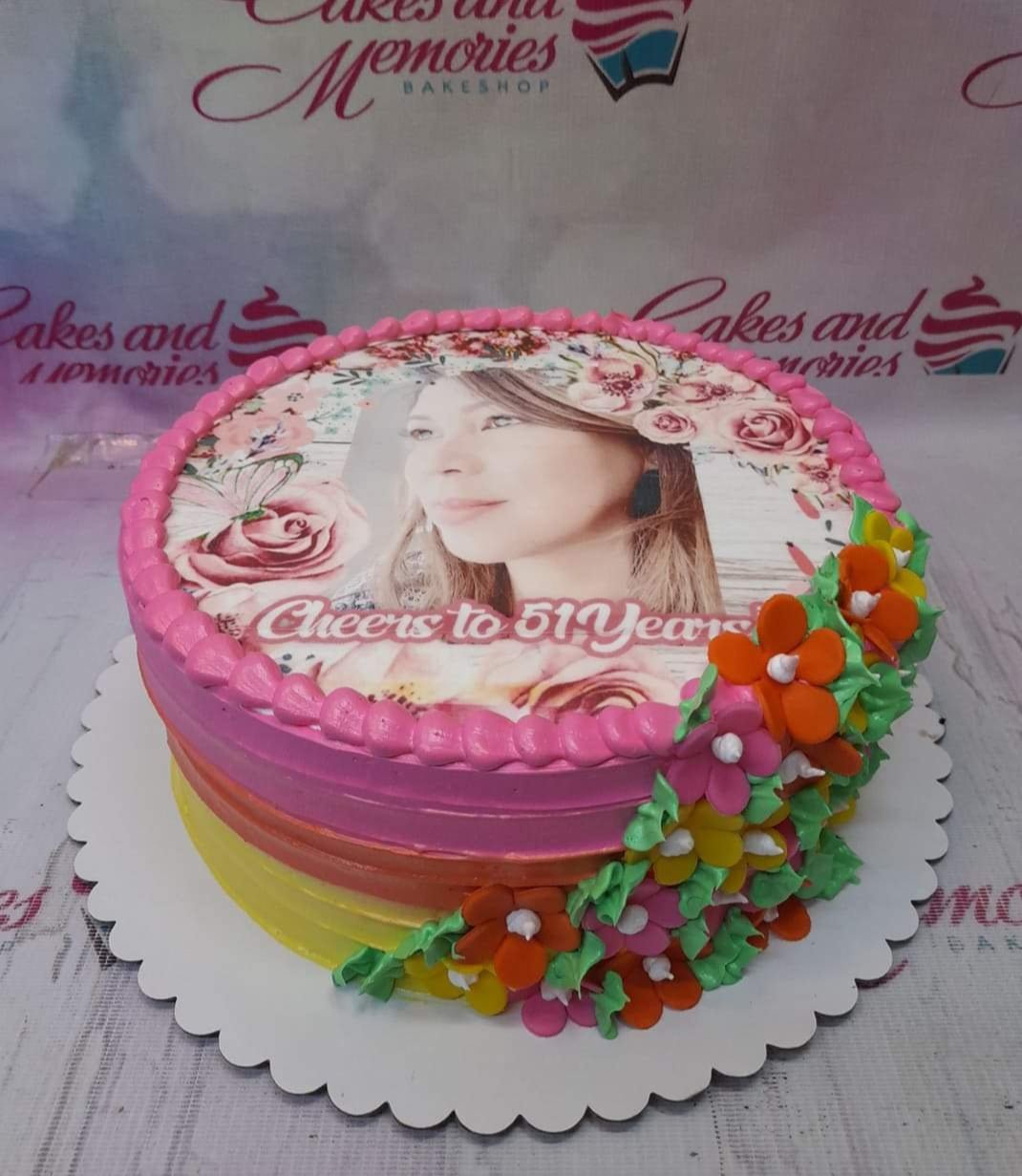 Round birthday cake with a large edible photo print of a woman, pink icing borders, and colorful icing flowers on the side.